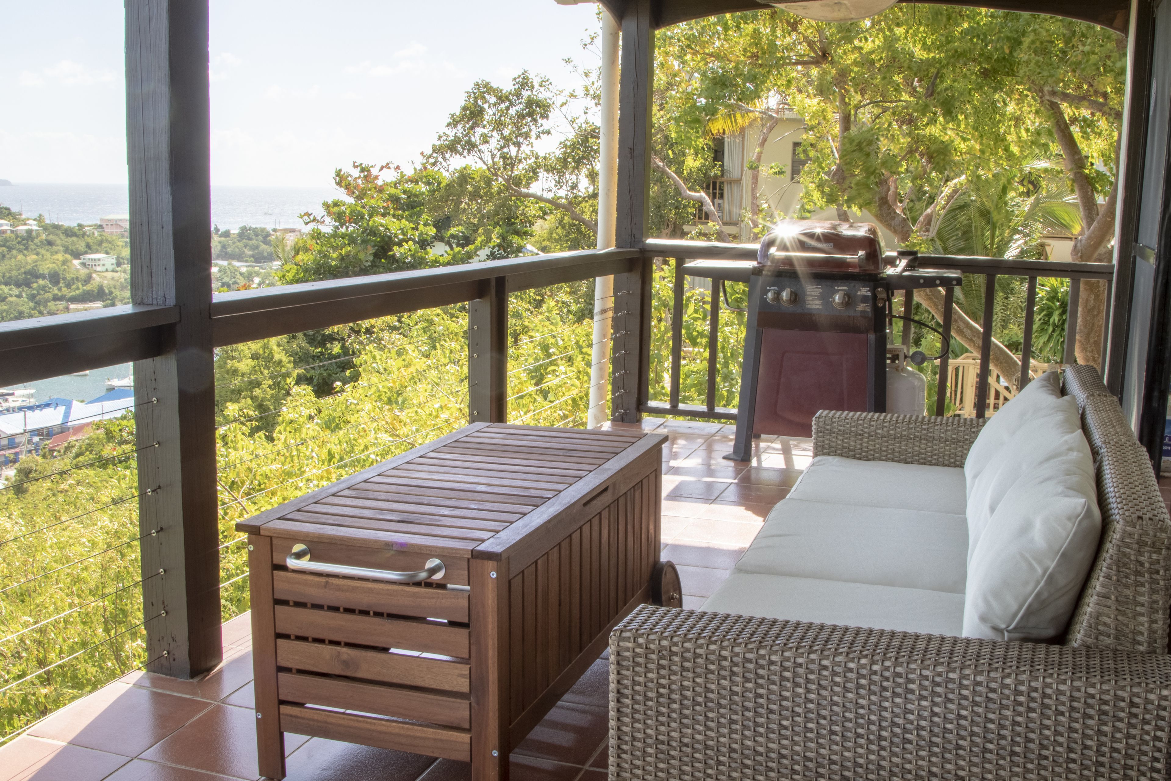 A balcony with a wooden table and wicker sofa, overlooking a lush green landscape and a body of water.