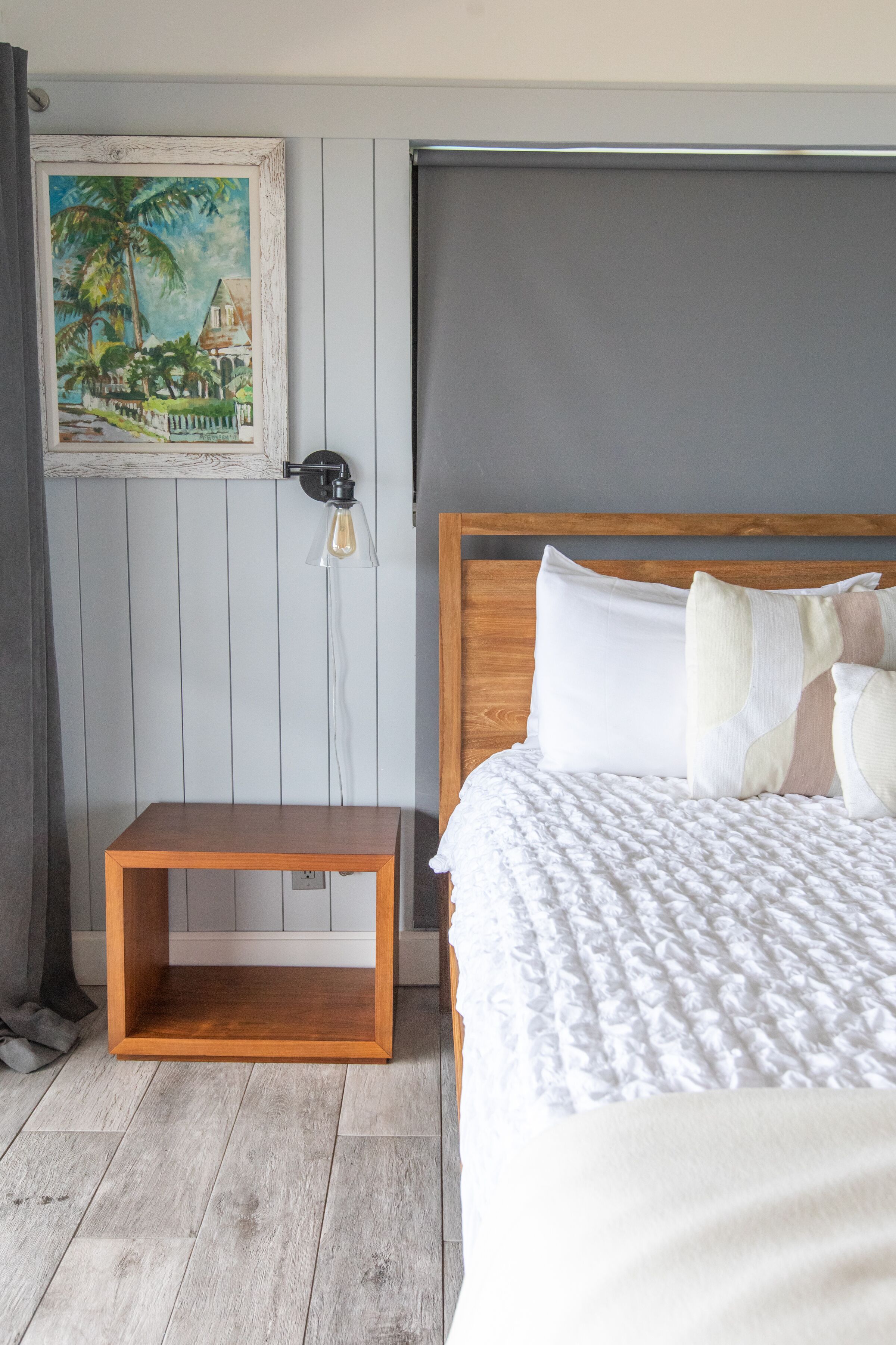 Een slaapkamer met een houten bed, een klein nachtkastje van hout, een wandlamp en een lijst met daarin een tropisch tafereel.