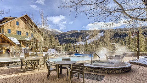 Una cabaña de madera con jacuzzi y zona de estar exterior, rodeada de montañas nevadas.