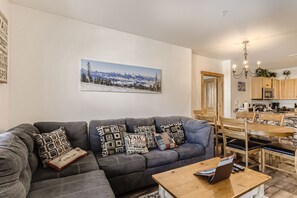 Uma sala de estar moderna com um sofá modular cinza, uma mesa de centro de madeira e uma área de jantar com cadeiras de madeira e uma mesa de mármore.