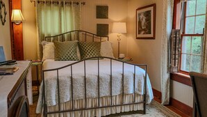 A bedroom with a metal bed frame, a bedside table with a lamp, a window with curtains, and a wall with framed artwork. - Romantic Inn suite with Air Jet Jacuzzi & private bath (Logan)