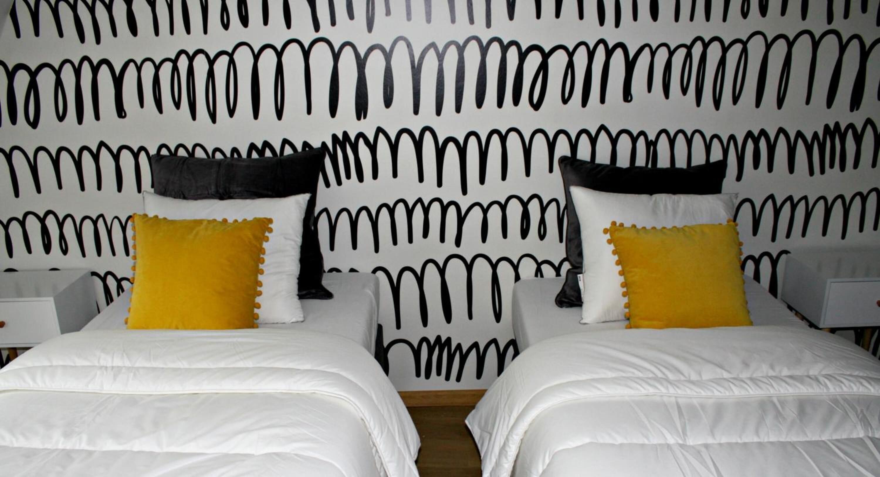 A hotel room with two beds, each with white bedding and yellow pillows. The room features a unique black and white abstract wall design.