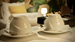 Ein ordentlich gedeckter Tisch mit zwei weißen Tassen und Untertellern, einem Löffel und einer kleinen Schale, vor dem Hintergrund eines Bettes mit einem Leopardenmuster-Kopfstück und einem Nachttisch mit einer Lampe.