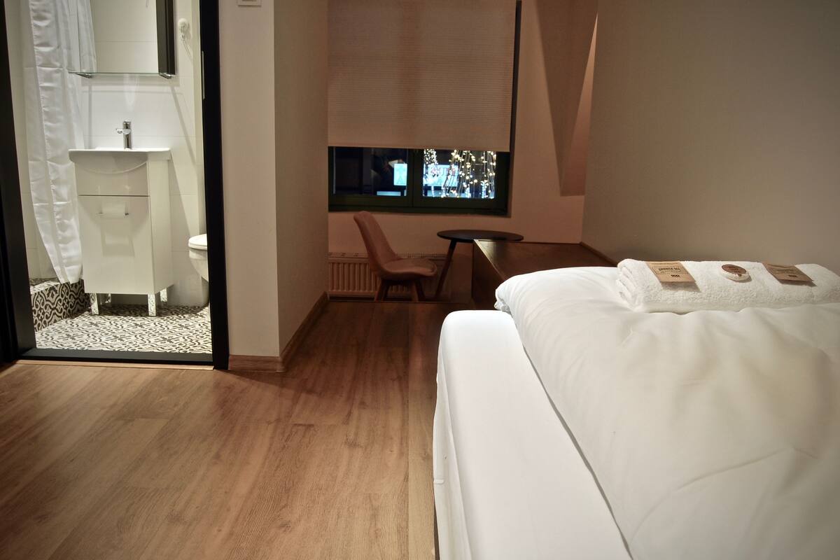 A hotel room with a bed, a chair, a small table, a window with a view, and a bathroom with a sink and toilet.