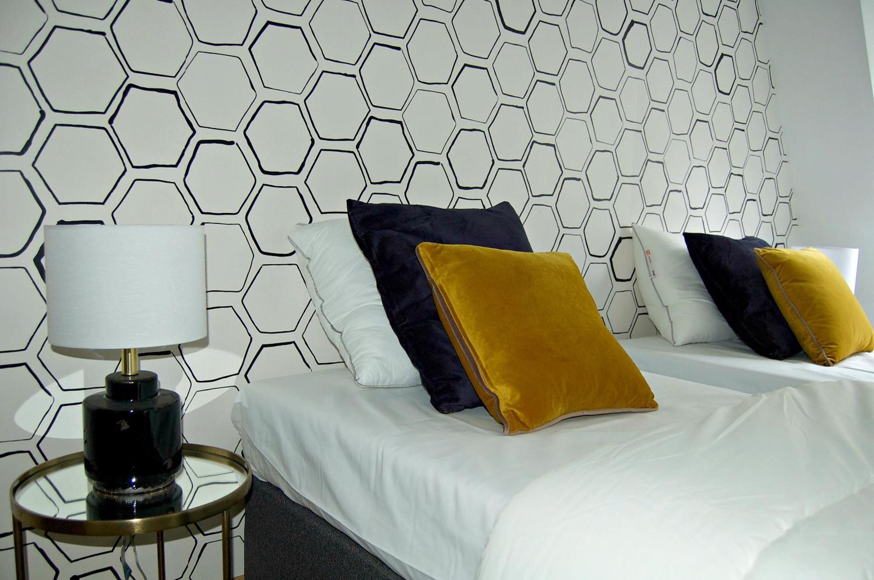 A hotel room with a modern hexagonal wall design, a round bedside table with a lamp, and two beds with white bedding and contrasting pillows.
