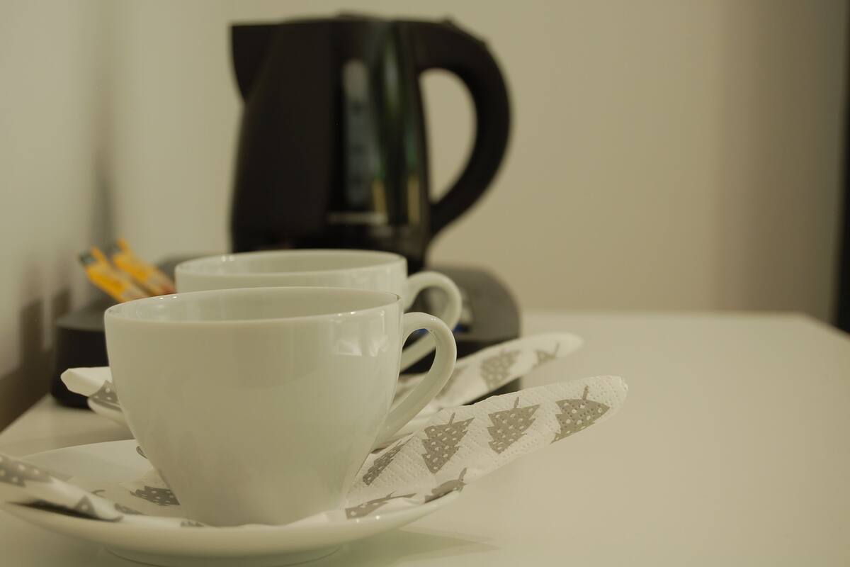 Two white cups on a saucer with a patterned napkin, a black coffee maker, and a spoon in the background.