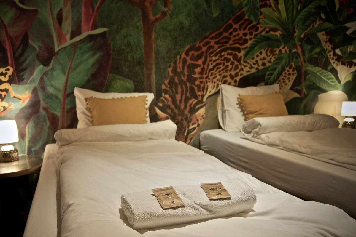 A hotel room with two beds, each with white linens and a pair of towels. There are two nightstands with lamps, and a large mural of a jungle scene on the wall behind the beds.
