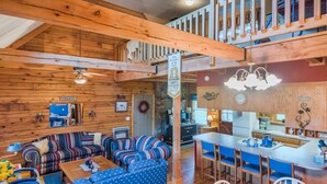 A cozy cabin interior with a wooden ceiling, a dining area with blue chairs, a kitchen with a white countertop, and a living room with striped sofas.