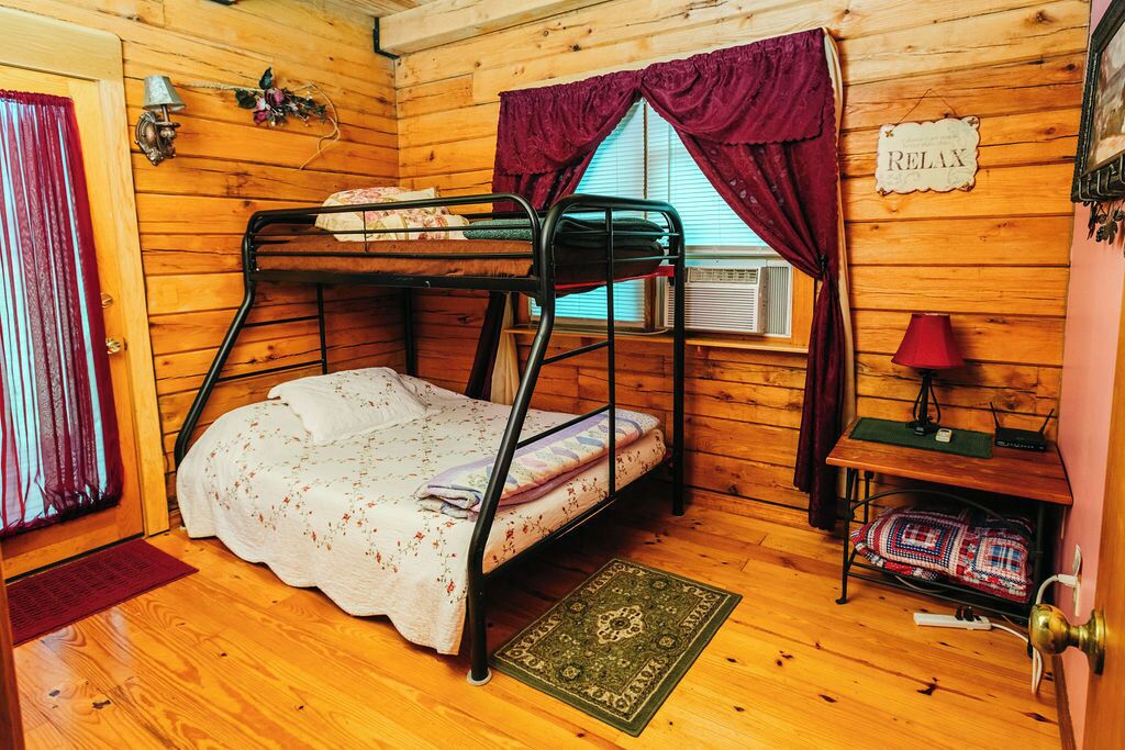 A cozy cabin room with a bunk bed, a small table with a lamp, a sign that says "RELAX", and a window with curtains.