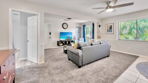 A spacious living room with a grey sofa, a ceiling fan, a large window with a view of greenery, and a clock on the wall. - Awesome Location! ☀Unlimited Sunsets Included☀ (Indian Rocks Beach)
