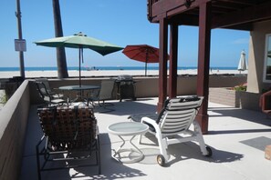 A beachside patio with outdoor seating, including wicker chairs and a white lounge chair, under a pergola with umbrellas. - Nothing beats Ocean Front (San Diego)