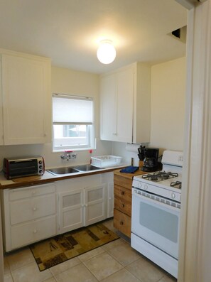 A compact kitchen with white cabinets, a toaster, a sink, a stove, and a window with blinds. - Nothing beats Ocean Front (San Diego)