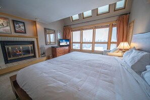 Une chambre à coucher avec un grand lit, une cheminée, un téléviseur et une vue sur la ville grâce à de grandes fenêtres.