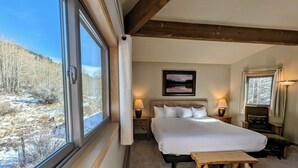 Une chambre avec un grand lit, deux tables de chevet avec des lampes, un banc en bois et une fenêtre donnant sur un paysage enneigé.