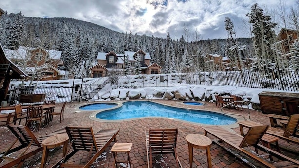A pool area with lounge chairs and a wooden deck, surrounded by snow-covered trees and buildings. - Settlers Creek 6514 (Keystone)