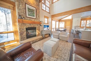 A spacious living room with a stone fireplace, leather sofas, and a view of the kitchen and dining area through large windows. - Settlers Creek 6565 (Keystone)
