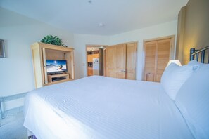 A bedroom with a large bed, a TV on a wooden stand, wooden wardrobes, and a kitchen visible through an open door. - Silver Mill 8255 (Keystone)