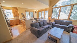 A spacious living area with a large gray sofa, a wooden coffee table, and a dining table with chairs. There is a kitchen area with wooden cabinets and a window overlooking a forested area. - Ironwood 2986 by SummitCove Vacation Lodging (Keystone)