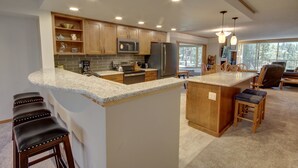 A modern kitchen with granite countertops, wooden cabinets, and stainless steel appliances. There is a large island with stools and a dining area with a view of the outdoors. - Pines 2086 (Keystone)