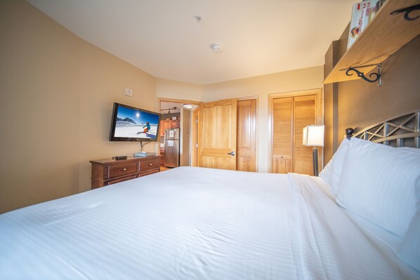 A hotel room with a large bed, wooden wardrobe, a television mounted on the wall, and a bedside table with a lamp. - Silver Mill 8283 (Keystone)