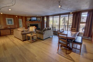 A spacious living room with a fireplace, wooden furniture, and large windows offering a view of the outdoors. - Pines 2074 (Keystone)