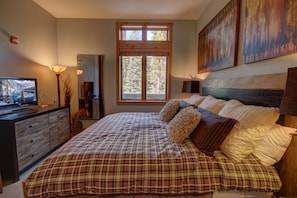 Un dormitorio con una cama grande, un televisor en una cómoda y una ventana con vista a árboles.