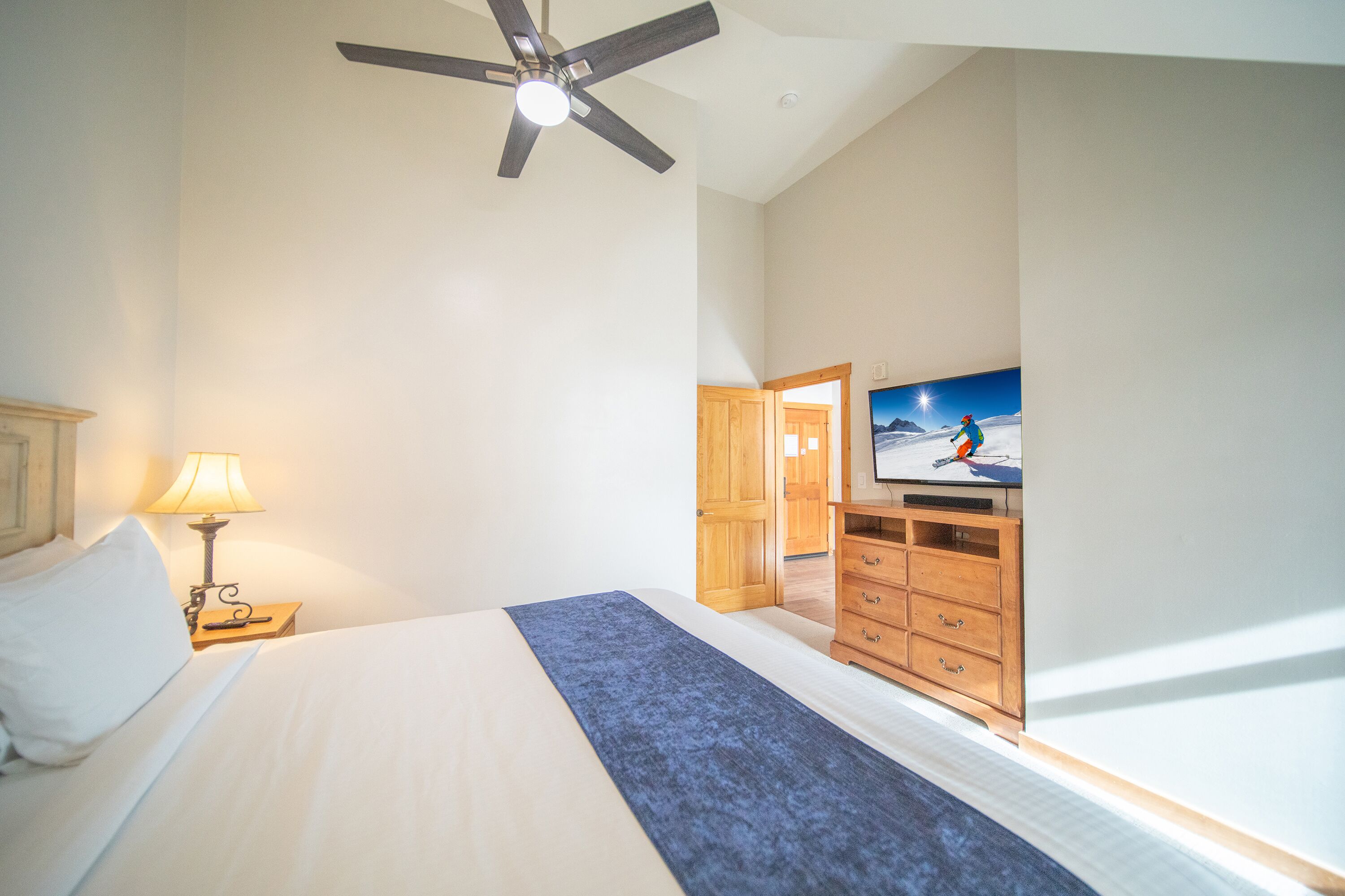 A hotel room with a bed, a ceiling fan, a TV, wooden furniture, and a view of the outdoors.
