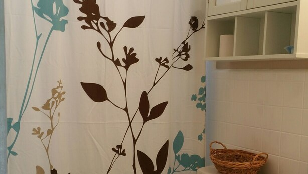 Une salle de bain avec un rideau de douche orné d’un motif floral, des toilettes, un panier en osier posé sur le lavabo et un meuble au-dessus du lavabo.