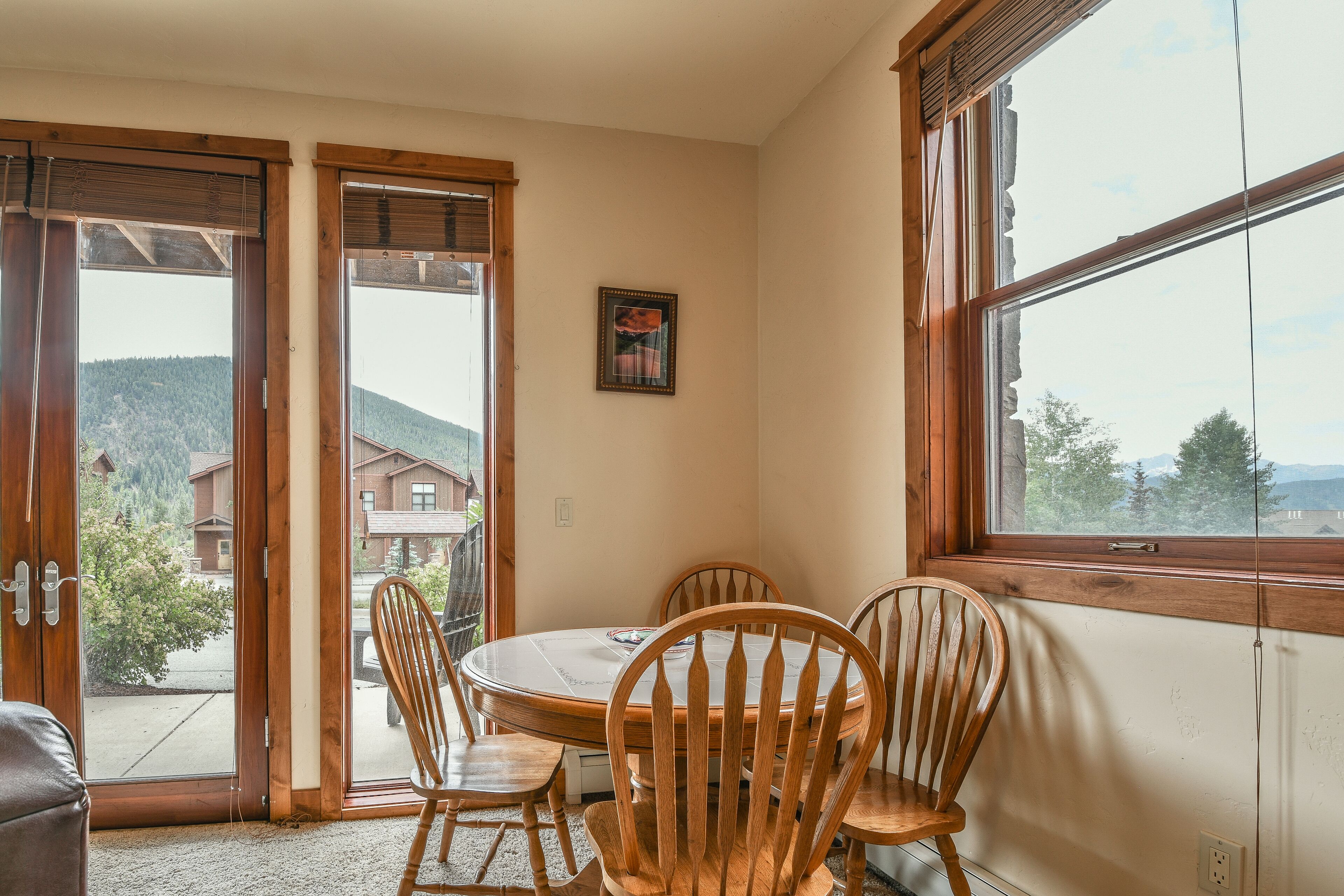 A dining area with a round table and wooden chairs, a window with a view of houses and trees, and a door leading outside.