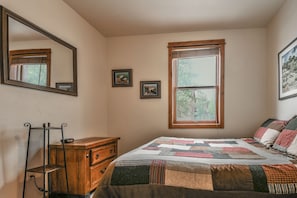 Een slaapkamer met een houten dressoir, een gewatteerde spreideken, een spiegel en een raam met uitzicht op bomen.