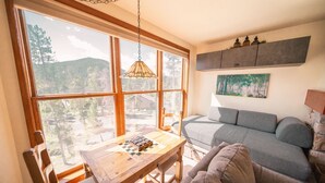 Una sala de estar acogedora con una mesa de madera, un sofá gris y un ventanal con vistas a un bosque.