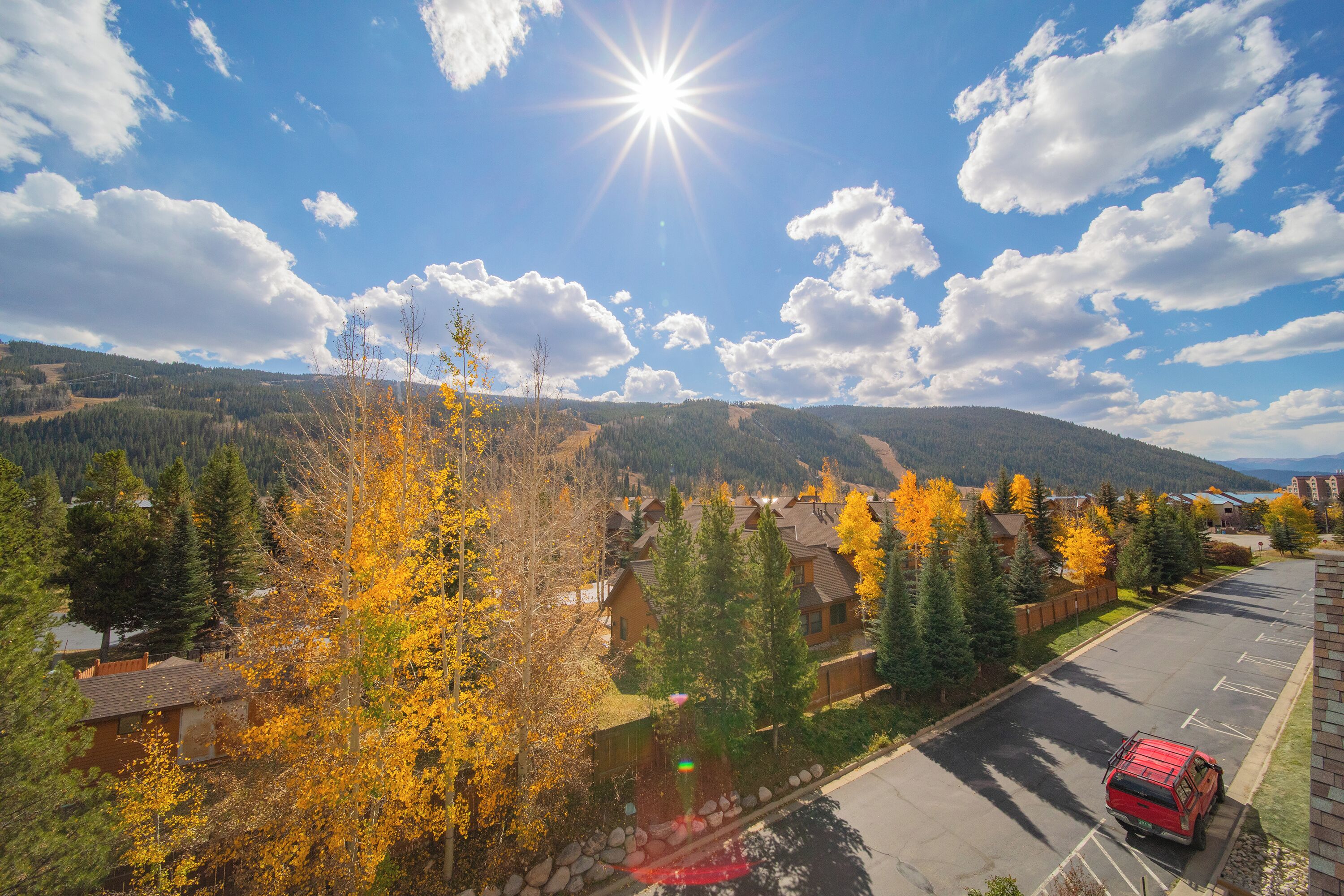 A parking lot with a red vehicle, surrounded by autumn-colored trees and houses with a mountain backdrop.