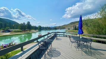 A wooden deck with outdoor seating overlooking a river, surrounded by greenery and buildings.