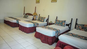 A hotel room with four single beds, each with a floral bedspread and a wooden headboard. There is a nightstand with a lamp and a telephone on each bed.