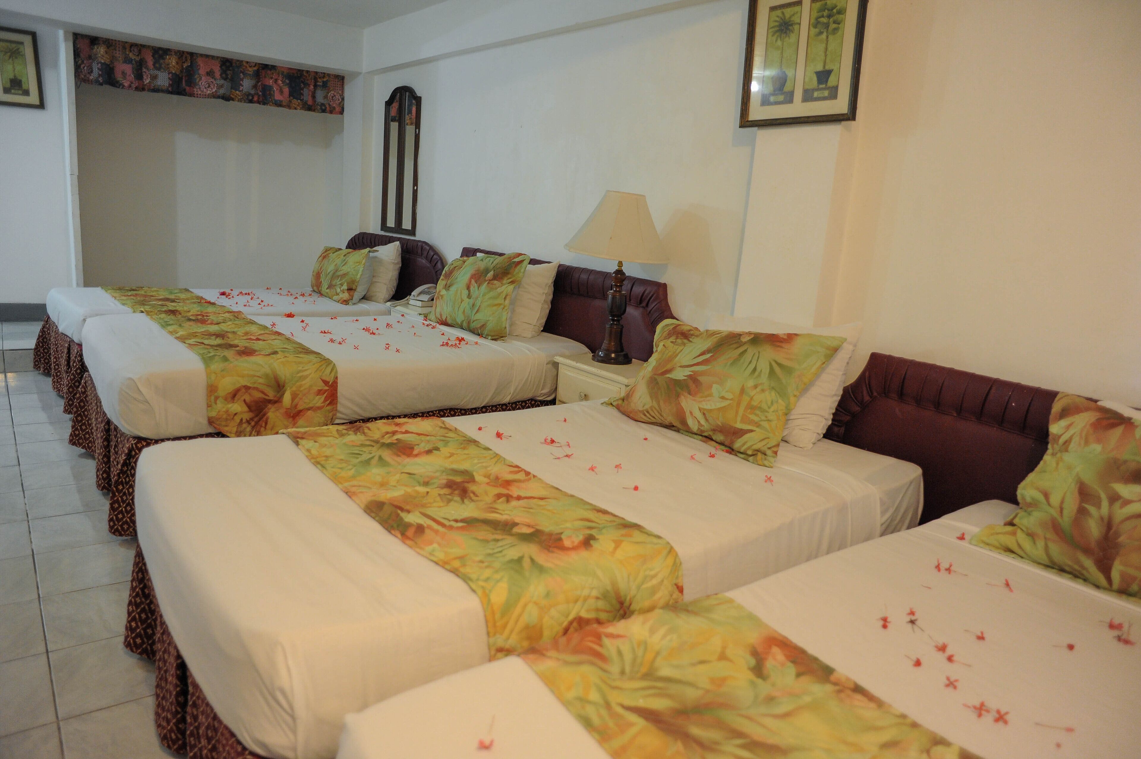 A hotel room with four beds arranged in a row, each with floral bedspreads and white linens. There is a large mirror on the wall and a lamp on the nightstand.