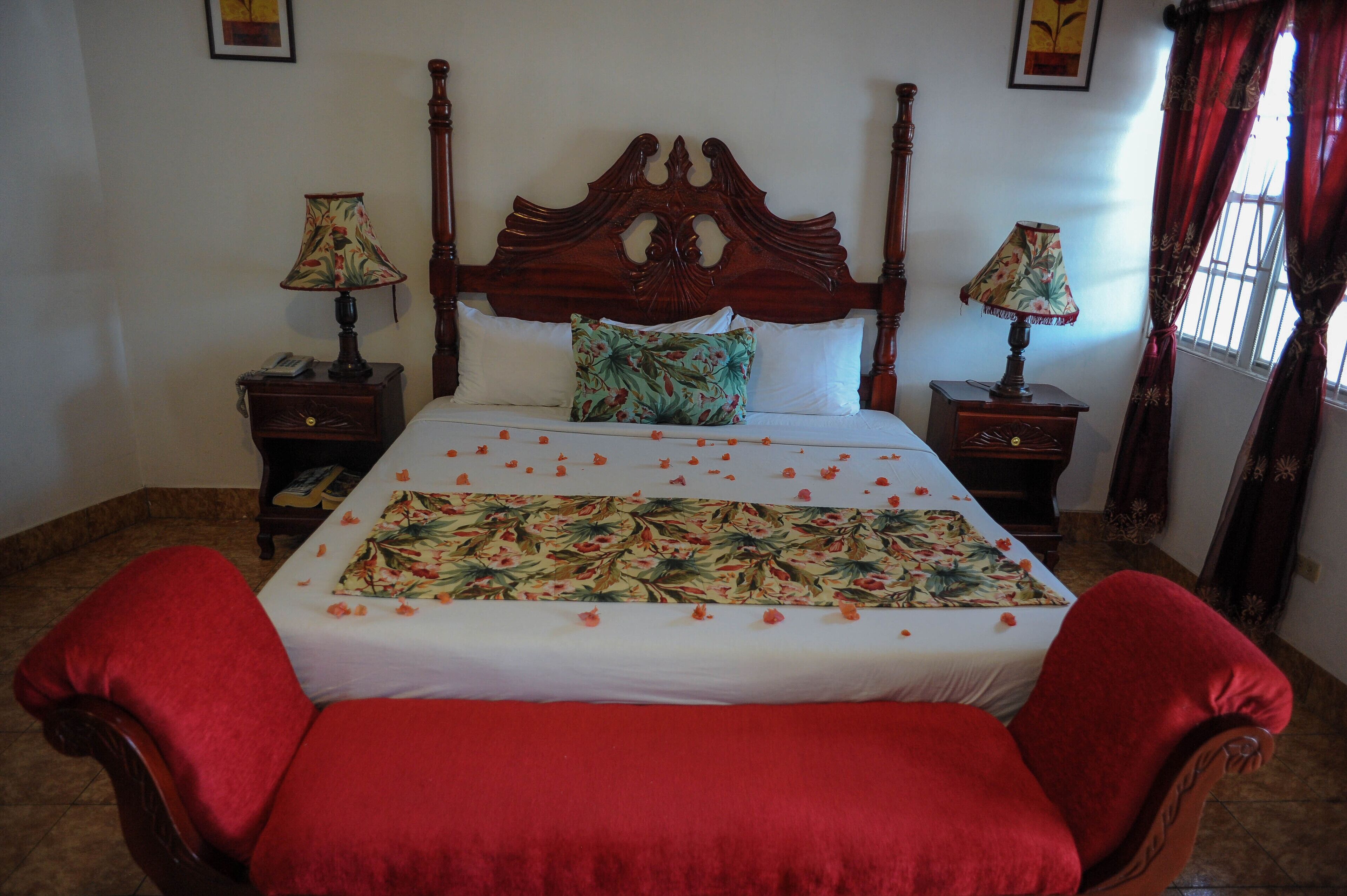 Una cama con colcha estampada, dos mesitas de noche con lámparas, un cabecero de madera y sillones tapizados en rojo.