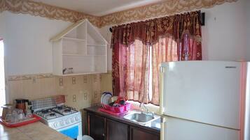Una cocina con estufa a gas, pileta y refrigerador. Hay una ventana con cortinas y un estante sobre la estufa.