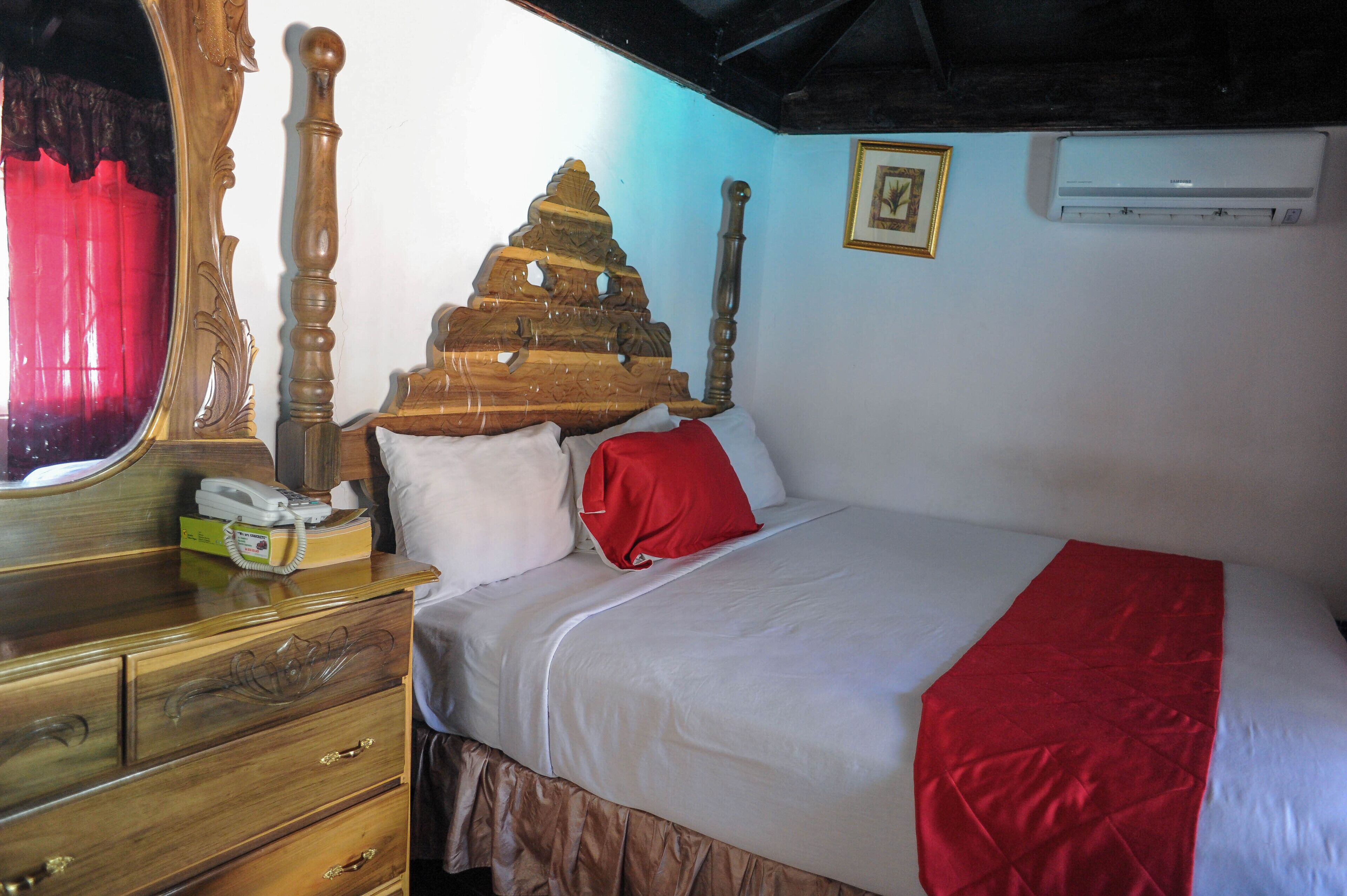 Une chambre à coucher avec un lit en bois, un téléphone sur la table de chevet, un miroir et un oreiller rouge.