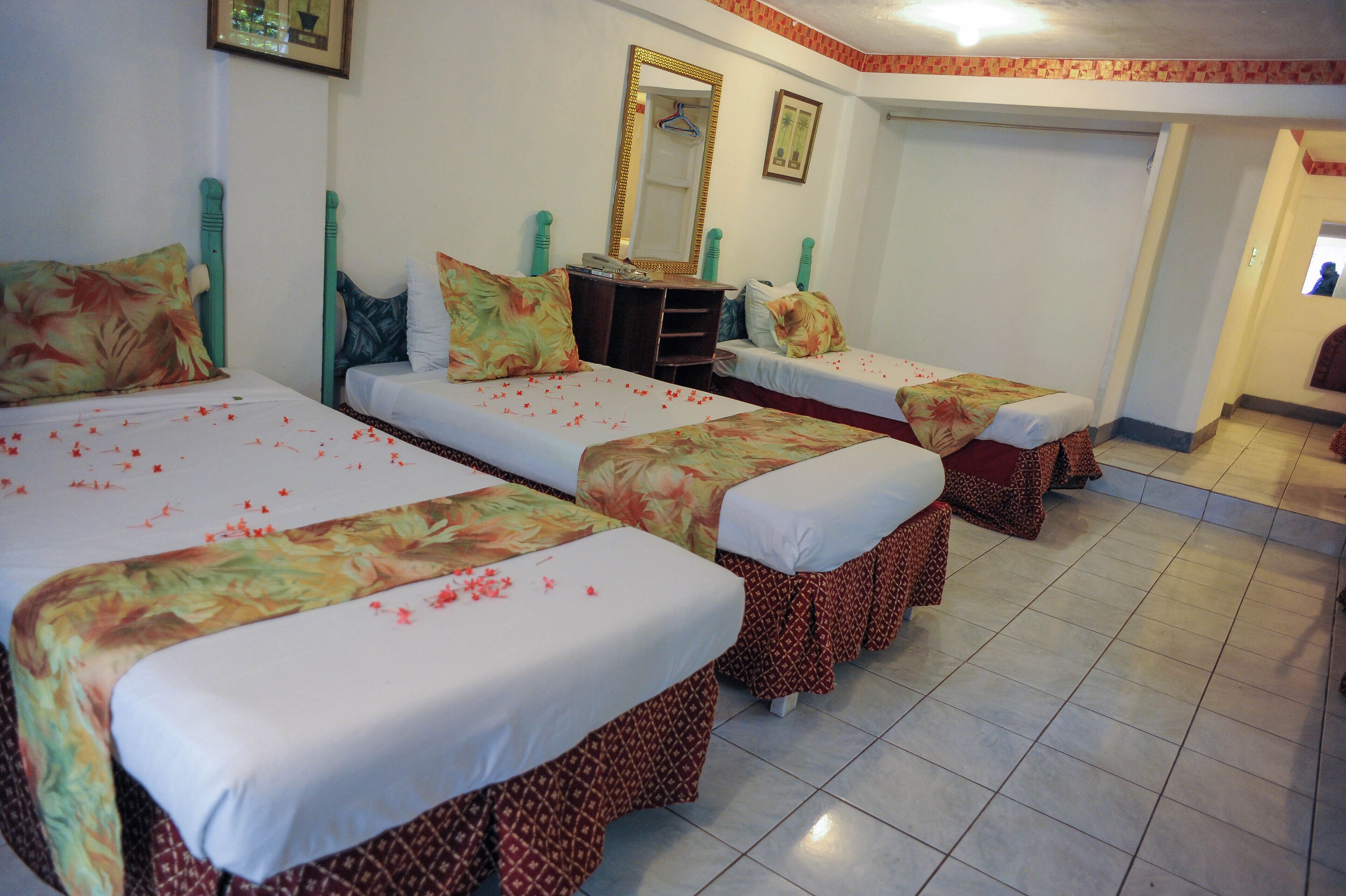 A hotel room with three single beds, each with a floral bedspread and a white pillow. There is a dresser with a mirror above it, and a door leading to another room.