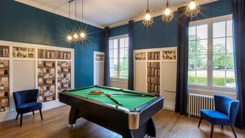Une table de billard dans une pièce dotée d’un plancher en bois, de bibliothèques et offrant une vue sur la verdure à l’extérieur.