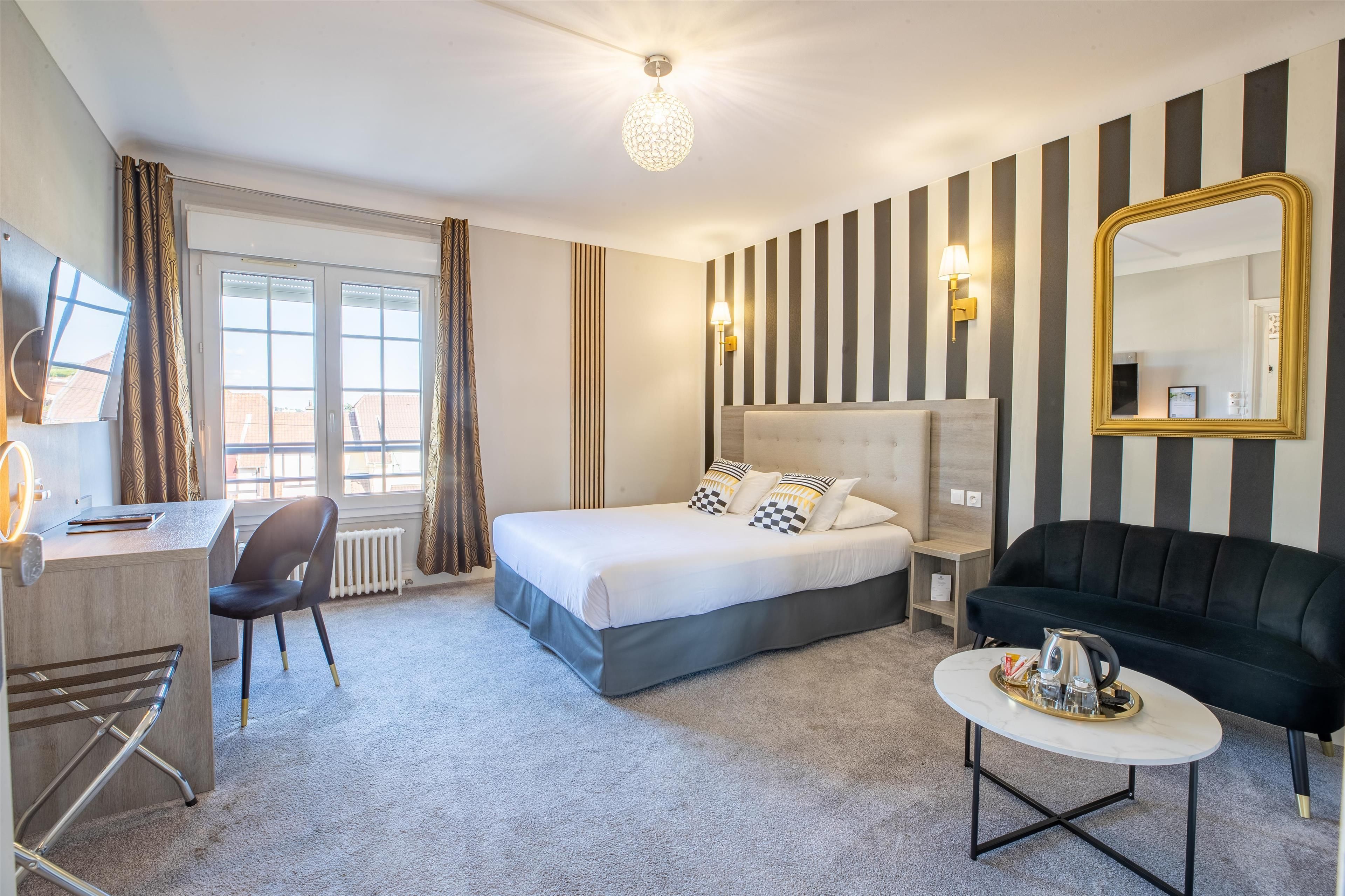 A hotel room with a bed, a desk, a chair, a black sofa, a small table with a tray, and a large mirror.