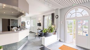 A modern hotel lobby with a reception desk, seating area, and large windows offering views of the outside. - Sure Hotel by Best Western Port Jerome - Le Havre (Port-Jérôme-sur-Seine)