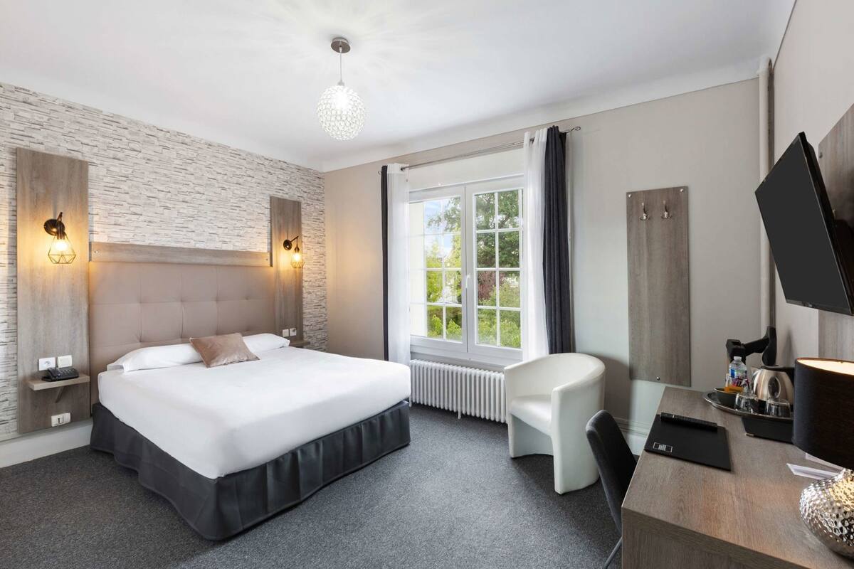Une chambre d’hôtel comprenant un lit, des tables de chevet, un bureau avec une chaise, une télévision et une fenêtre avec des rideaux.