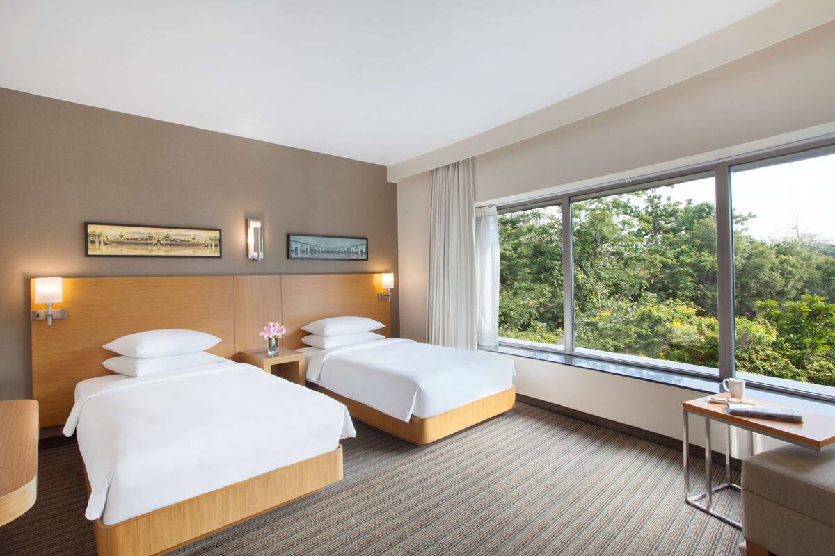 A hotel room with two beds, a desk, a window with a view of greenery, and wall-mounted artwork.