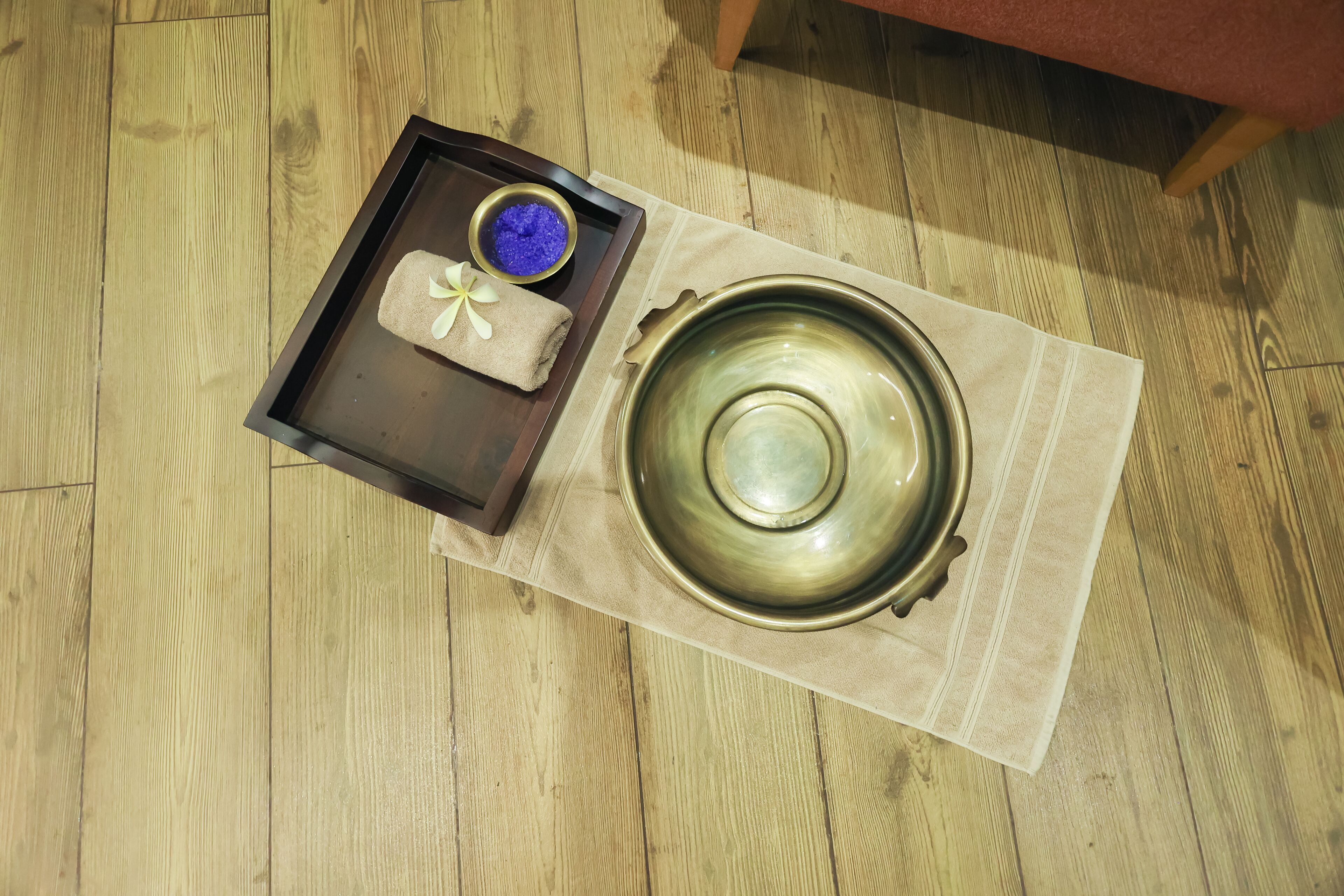 A wooden floor with a tray containing a towel, a bowl of blue bath salts, and a ceramic bowl on a beige cloth.