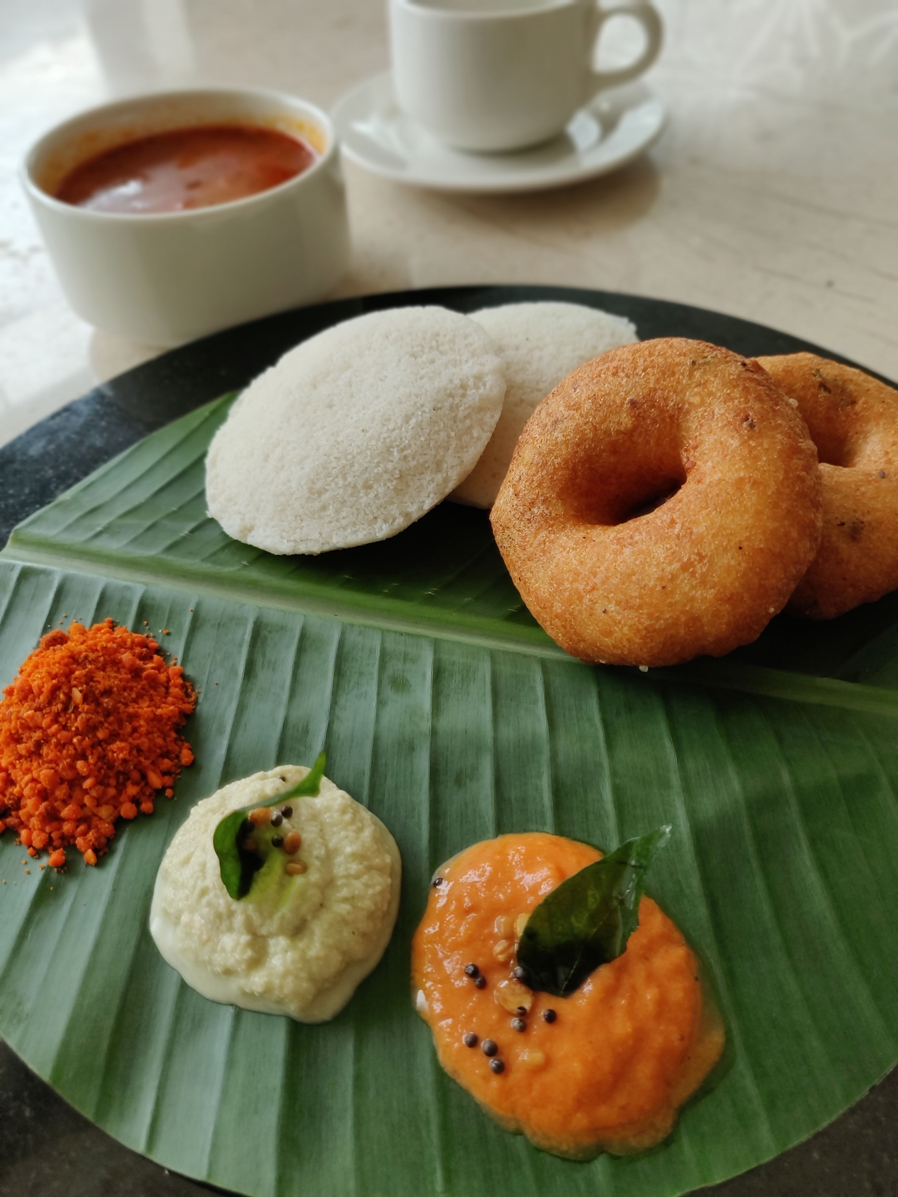 Un plato de comida con idlis, vadas y chutneys sobre una hoja de plátano.
