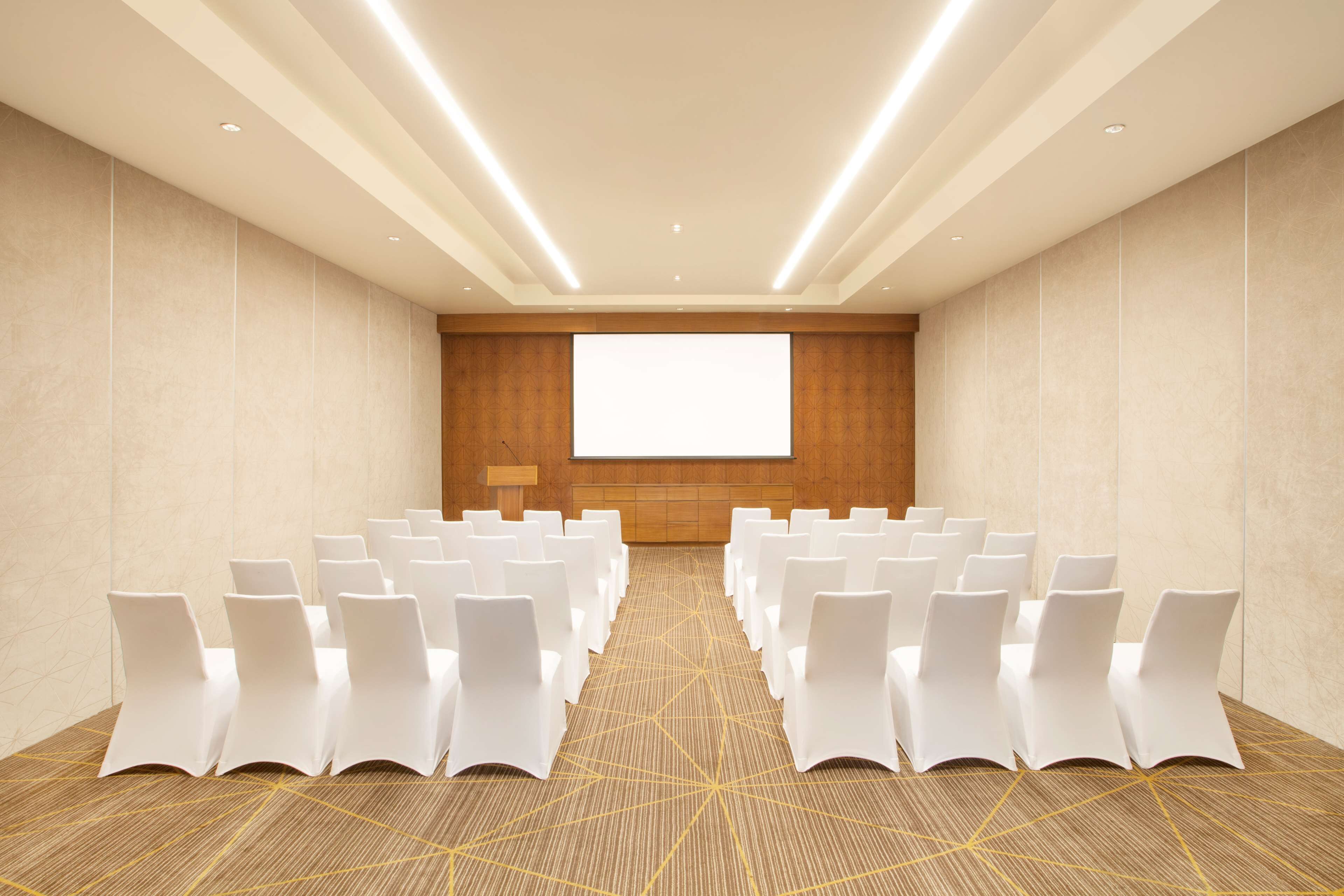 A conference room with rows of white chairs, a large projection screen, and a carpeted floor.