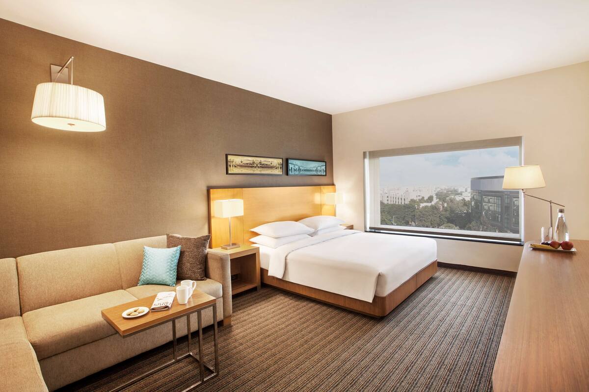 A hotel room with a bed, a sofa, a small table with a tray of apples, a lamp, and a window with a city view.