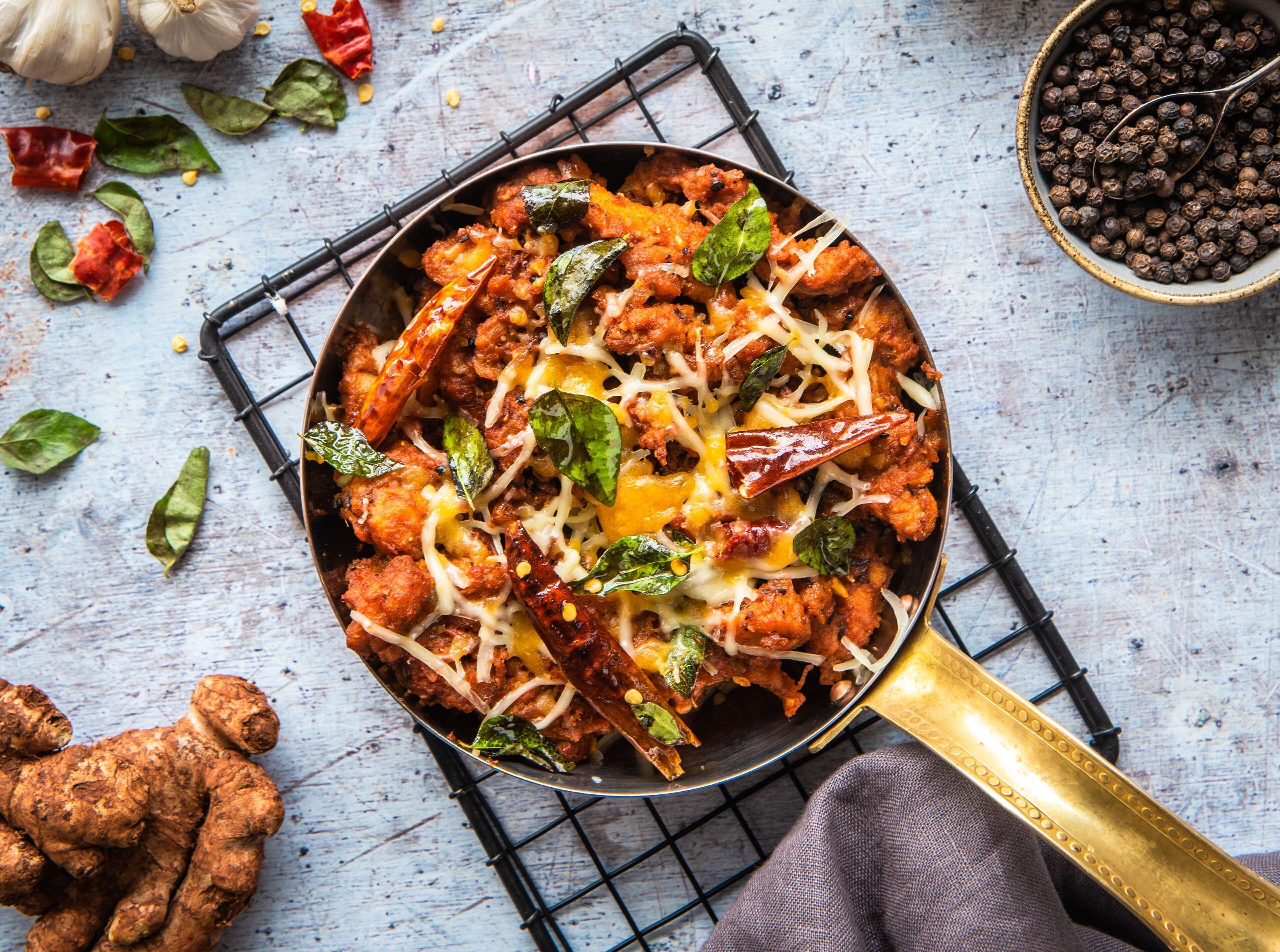 Una sartén con un plato que contiene carne, queso y hierbas, rodeado de ajos, chiles y un tazón de granos de pimienta negra sobre una superficie de madera.