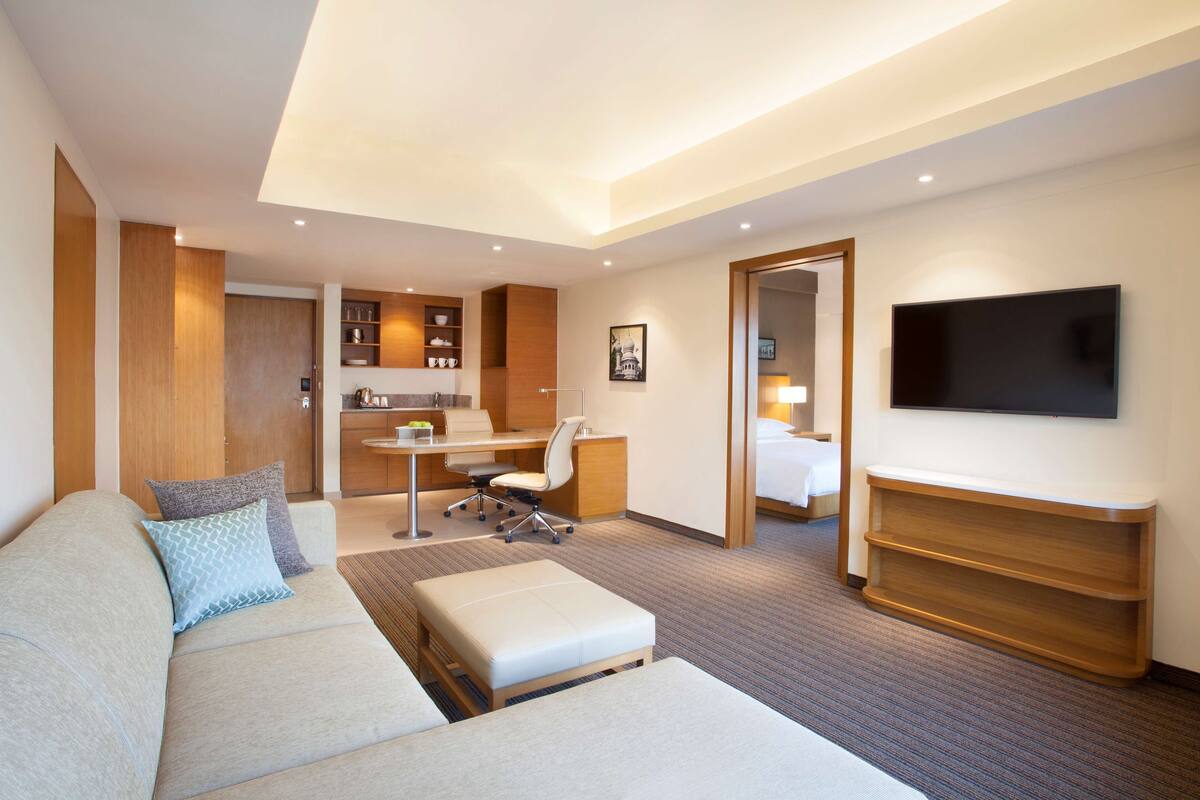 A modern hotel room with a sofa, a desk, a chair, a TV, and a kitchenette.